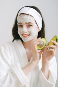 девушка в белом халате с маской из авокадо и их плодами в руках