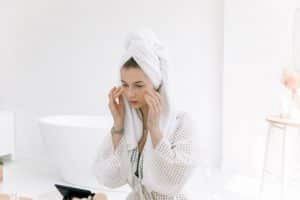девушка в халате и с полотенцем на голове ухаживает за лицом в ванной комнате перед зеркалом