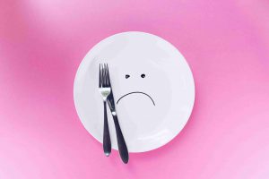 белая тарелка, на которой нарисована грустная мордочка, вилка, нож и розовый фон сзади